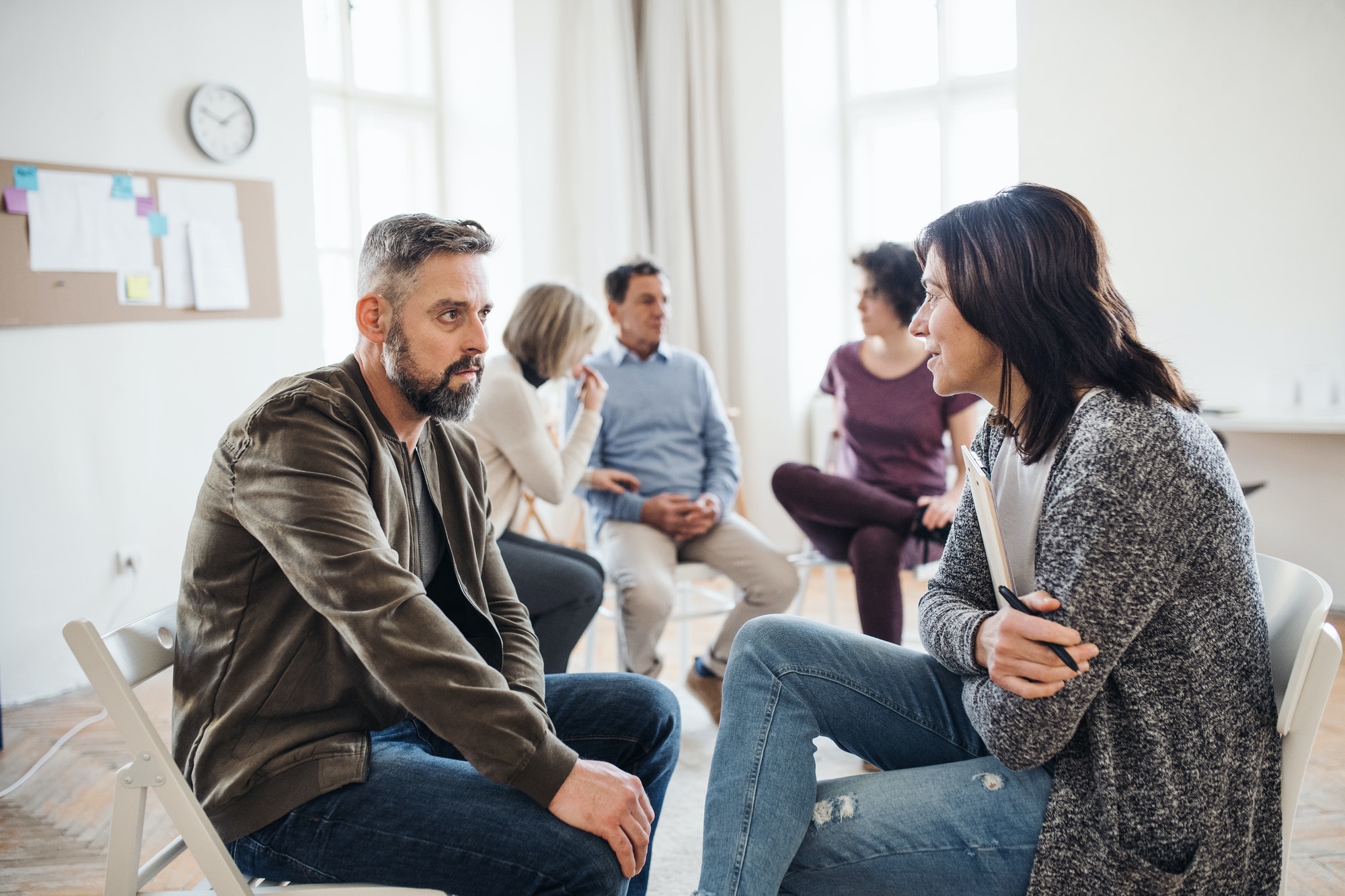 senior-counselor-with-clipboard-talking-to-a-man-during-group-therapy-.jpg